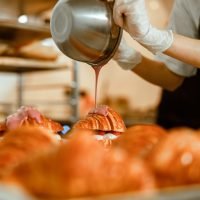 Confectioner pours pink icing onto tasty croissant from metal bowl on counter